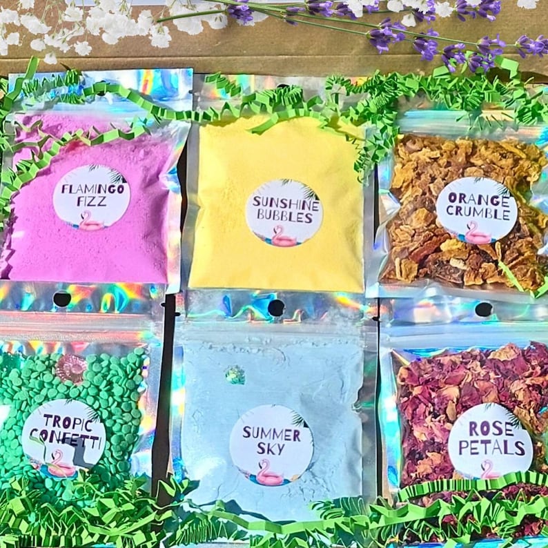 May include: A collection of colorful bath products in clear pouches, including pink "Flamingo Fizz", yellow "Sunshine Bubbles", and orange "Orange Crumble". Other pouches contain green confetti, blue "Summer Sky", and rose petals. Each pouch has a circular label with a flamingo.