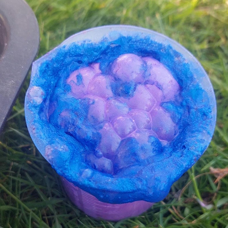 May include: A close-up shot of a purple and blue bath bomb in a small, clear container. The bath bomb has a bubbly texture and is surrounded by a vibrant blue color. The container is sitting on green grass.
