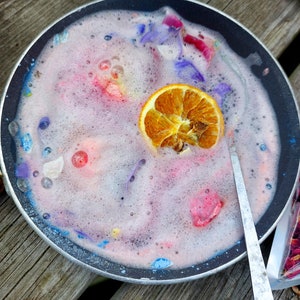 May include: A black pan filled with pink, bubbly liquid, colorful flower petals, and a dried orange slice. The liquid has a foamy texture, and a silver spoon rests on the side. The scene is set on a wooden surface, suggesting a spa or beauty product theme.