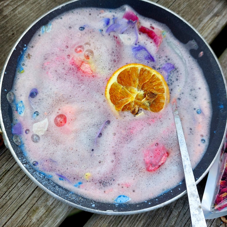 May include: A black pan filled with pink, bubbly liquid, colorful flower petals, and a dried orange slice. The liquid has a foamy texture, and a silver spoon rests on the side. The scene is set on a wooden surface, suggesting a spa or beauty product theme.