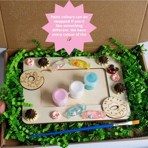 May include: A wooden picture frame kit with paint pots, a paintbrush, and colorful embellishments. The frame is decorated with donut and candy shapes. The text on the box reads "Paint colors can be swapped if you'd like something different. We have every color of the"