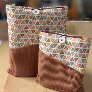 Peut inclure: Deux étuis à livres en tissu. La partie supérieure présente un motif géométrique de triangles dans des tons pêche, verts et dorés sur fond blanc. La moitié inférieure est en tissu uni côtelé de couleur rouille. Chaque étui a une fermeture à bouton blanc.