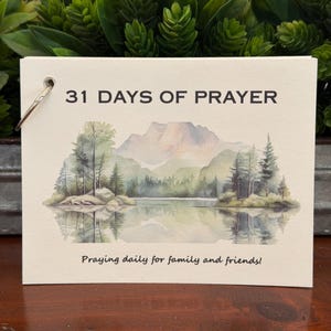 Puede incluir: Una tarjeta rectangular con un orificio para anilla metálica, con el texto 31 DAYS OF PRAYER en la parte superior y Praying daily for family and friends! en la inferior. El centro muestra un paisaje en acuarela de un lago, árboles y montañas.