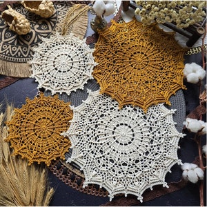 Op de afbeelding: Vier gehaakte doilies in wit en mosterdgeel. De doilies zijn gerangschikt op een donker oppervlak met tarwehalmen en wattenbolletjes.