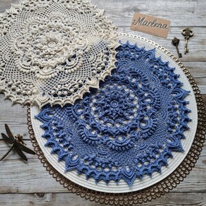 May include: Two crocheted doilies, one in white and one in blue, are displayed on a white base with a gold metal trim. The white doily is on top of the blue doily. The text "Marlena" is visible on a brown tag.