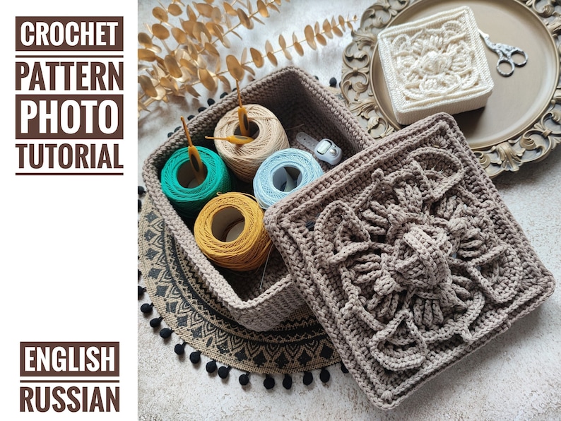 May include: A crocheted storage box with a lid, featuring a detailed floral design. The box is beige and the lid is brown. The box is filled with yarn balls in various colors, including green, blue, and yellow. The box is sitting on a black and white crocheted rug with black pom-poms.