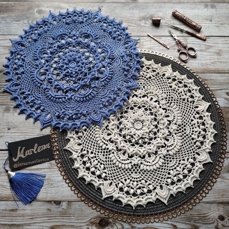 May include: Two crocheted doilies, one blue and one white, are displayed on a brown woven mat with a gold trim. The doilies are circular and feature intricate patterns. The blue doily has a tag with the text "Marlena @lenamasterica".