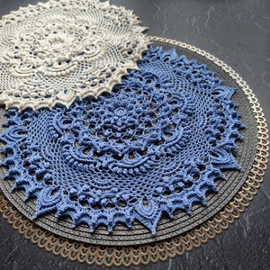 May include: Two crocheted doilies, one white and one blue, are layered on top of each other. The doilies are placed on a black and gold circular placemat.