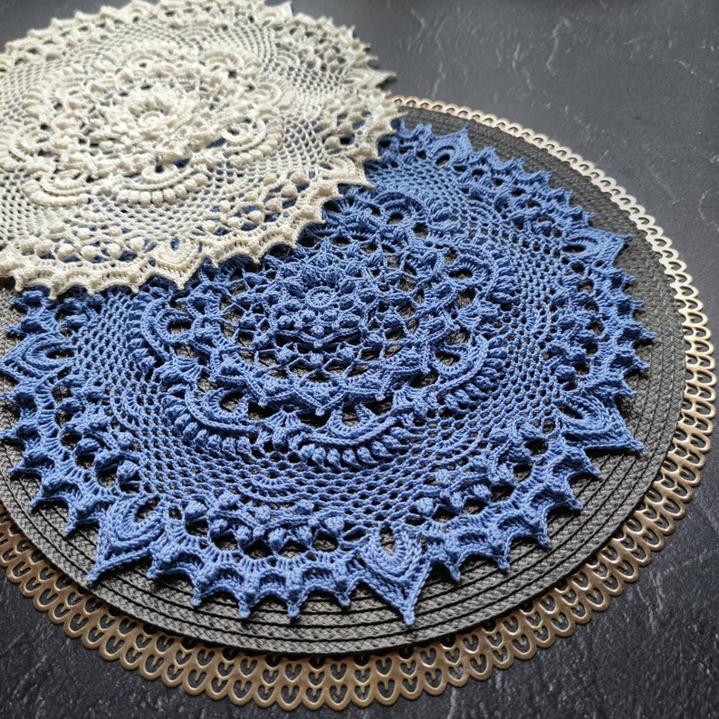May include: Two crocheted doilies, one white and one blue, are layered on top of each other. The doilies are placed on a black and gold circular placemat.