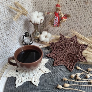 Puede incluir: Un posavasos de ganchillo marr&oacute;n con un dise&ntilde;o de estrella se encuentra sobre un posavasos de ganchillo gris y blanco. Una taza de caf&eacute; se encuentra encima de los posavasos.