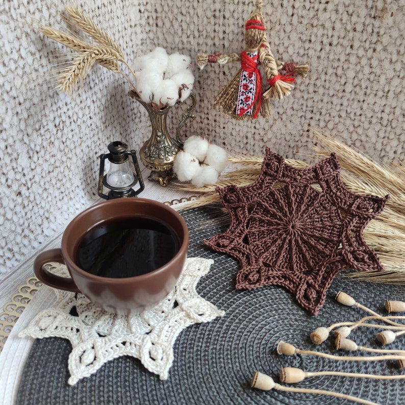 Puede incluir: Un posavasos de ganchillo marr&oacute;n con un dise&ntilde;o de estrella se encuentra sobre un posavasos de ganchillo gris y blanco. Una taza de caf&eacute; se encuentra encima de los posavasos.