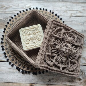 May include: A brown crocheted square box with a white crocheted square inside. The box has a decorative pattern and is sitting on a brown and black patterned surface.