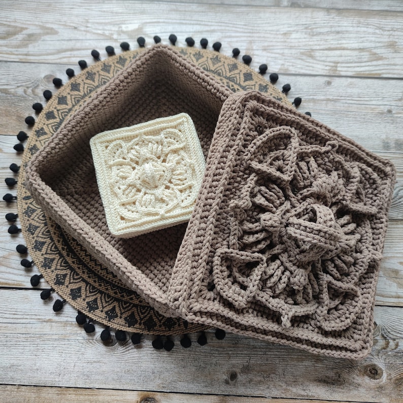 May include: A brown crocheted square box with a white crocheted square inside. The box has a decorative pattern and is sitting on a brown and black patterned surface.