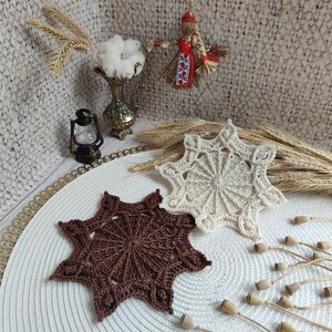 Puede incluir: Dos posavasos de estrella de ganchillo, uno marr&oacute;n y otro beige, sobre un mantel circular blanco y dorado.