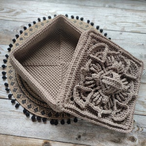 May include: A crocheted square box with a lid. The box is made of brown yarn and has a decorative pattern on the lid. The box is sitting on a brown and black woven mat.