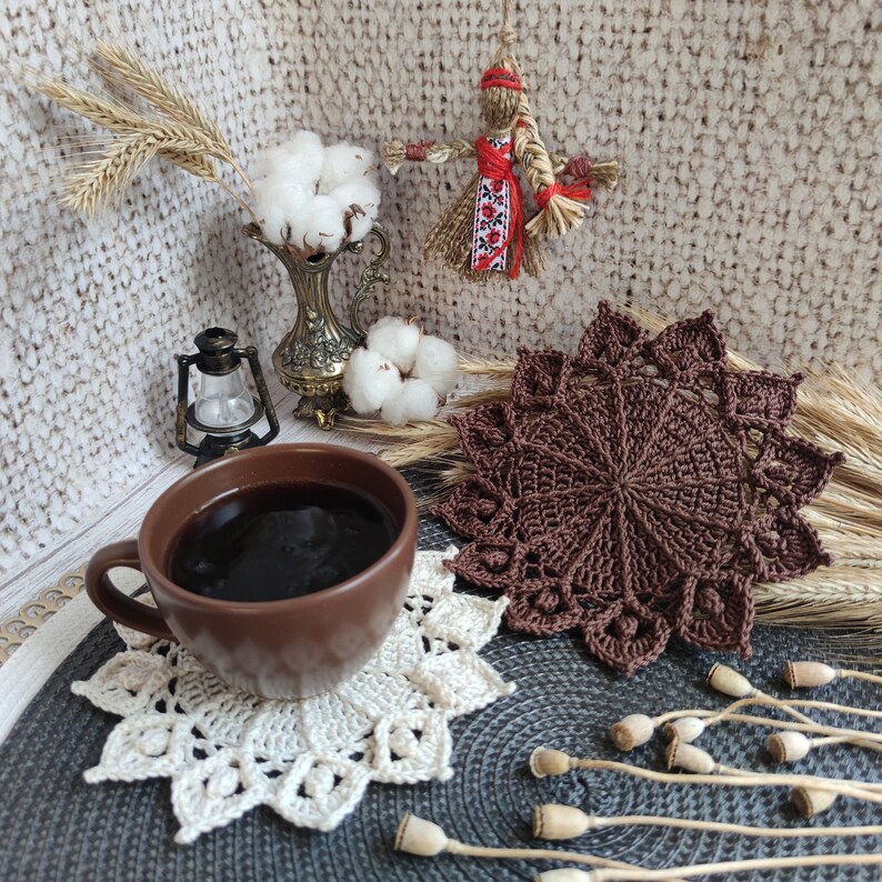 May include: A brown crocheted coaster with a white crocheted doily on top. A brown ceramic mug with coffee is sitting on the doily. The mug is next to a small, antique-looking lantern. There is a straw doll with red and white clothing in the background.