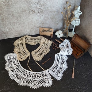 K&ouml;nnte beinhalten: Zwei handgefertigte H&auml;kelkragen in Creme und Wei&szlig;, pr&auml;sentiert auf einer dunklen Oberfl&auml;che. Die Kragen weisen komplizierte Spitzenmuster und filigrane Details auf. Ein kleines Etikett mit dem Namen "Amelie" ist sichtbar. Weitere Artikel sind Schere, N&auml;hzeug und eine dekorative Schaufensterpuppe.