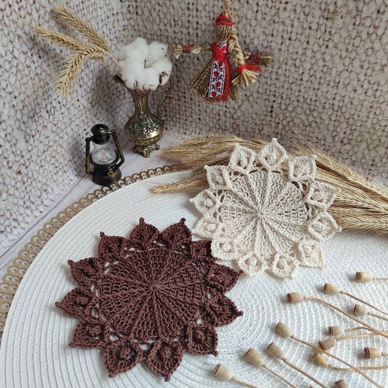 May include: Two crocheted doilies, one brown and one cream, on a white surface with dried flower pods.