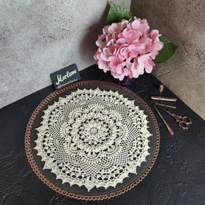 May include: A white crocheted doily with a delicate floral pattern, displayed on a copper-colored round tray. The doily is surrounded by a dark background.
