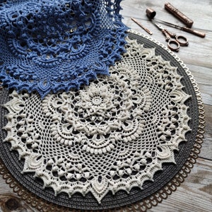 May include: Two crocheted doilies, one white and one blue, are displayed on a black and gold circular mat. The doilies are delicate and intricate, with a floral design.