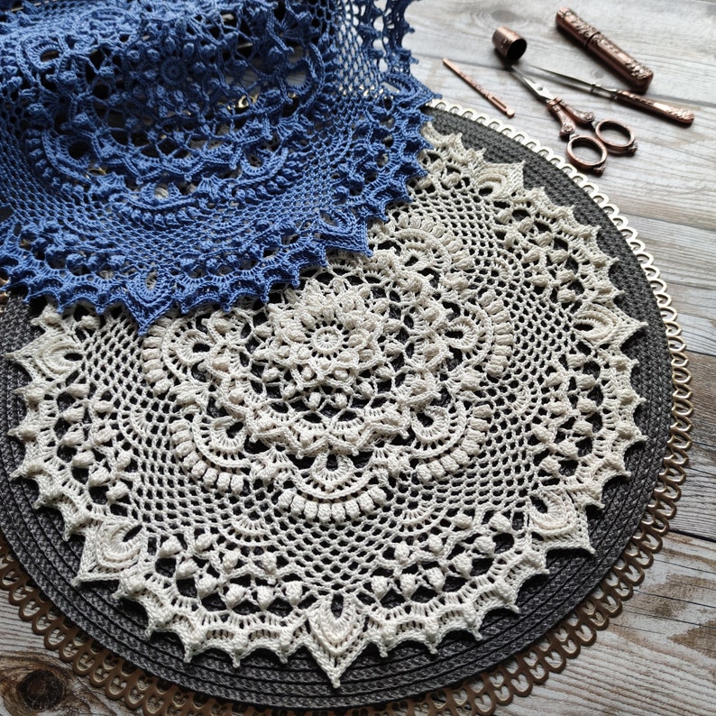 May include: Two crocheted doilies, one white and one blue, are displayed on a black and gold circular mat. The doilies are delicate and intricate, with a floral design.