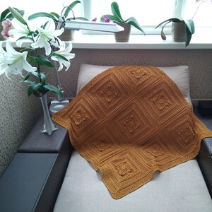May include: A mustard-colored, square-patterned blanket with floral designs is draped over a chair. The blanket's intricate details are highlighted by the warm color. White lilies in a vase and potted orchids are in the background.