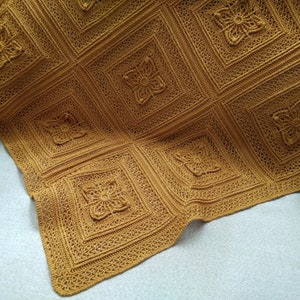 May include: Mustard-colored crocheted blanket with a square pattern. Each square features a floral design in the center. The blanket is made of a soft yarn and has a textured appearance. The blanket is folded over on a light gray surface.