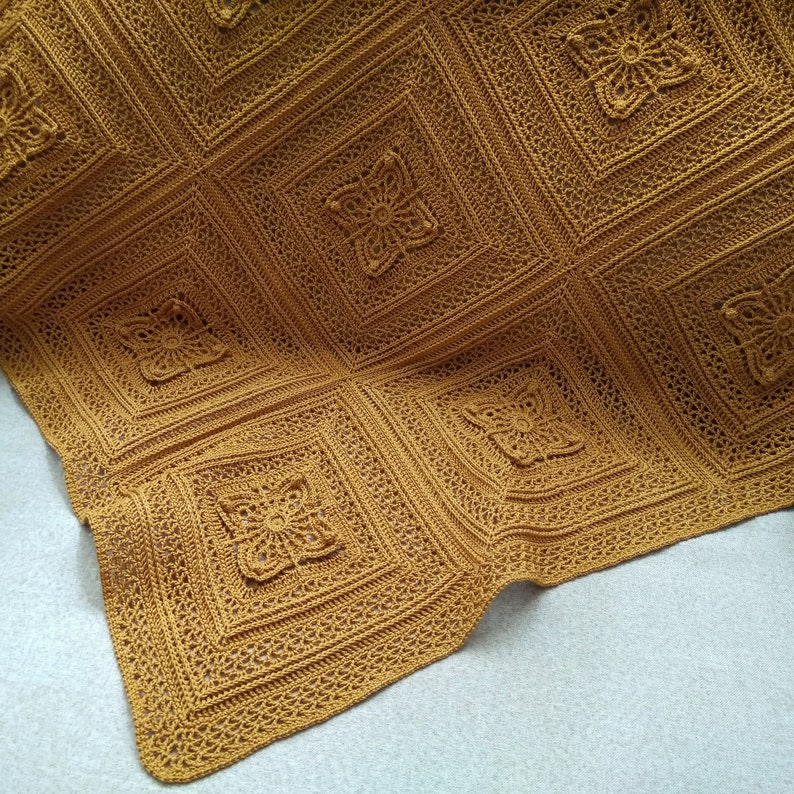 May include: Mustard-colored crocheted blanket with a square pattern. Each square features a floral design in the center. The blanket is made of a soft yarn and has a textured appearance. The blanket is folded over on a light gray surface.