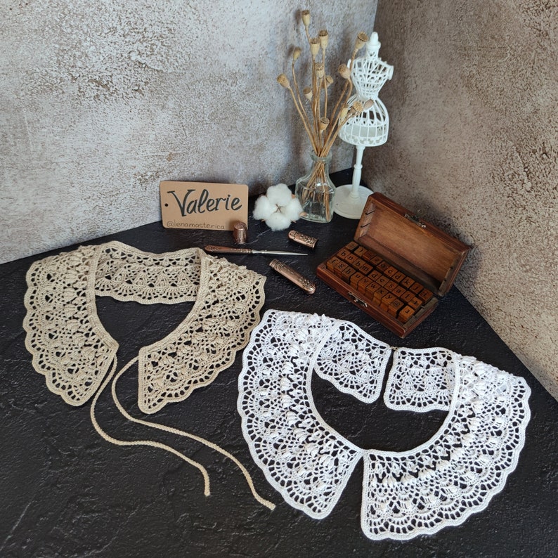 May include: Two crocheted collars, one beige and one white, with delicate lace patterns. The collars are displayed on a dark surface with a wooden box and other decorative items.