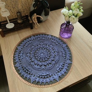 May include: A purple crochet doily with a delicate lace pattern sits on a wooden table with a copper-colored trim. The doily is a circular design with a central motif and intricate details.
