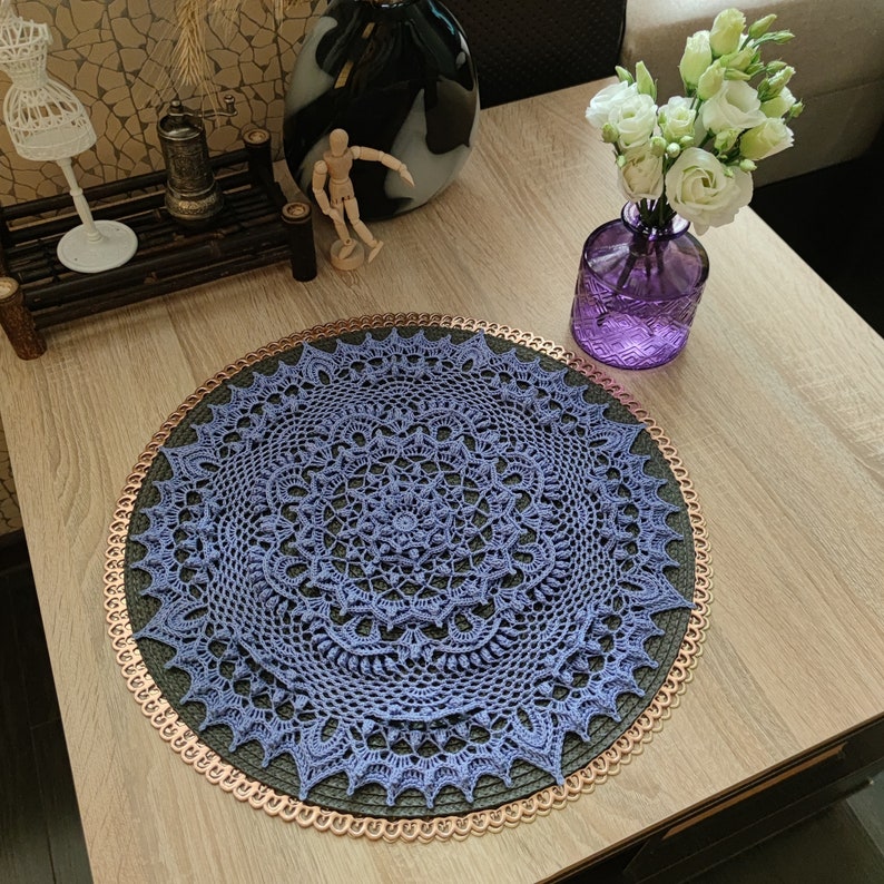 May include: A purple crochet doily with a delicate lace pattern sits on a wooden table with a copper-colored trim. The doily is a circular design with a central motif and intricate details.
