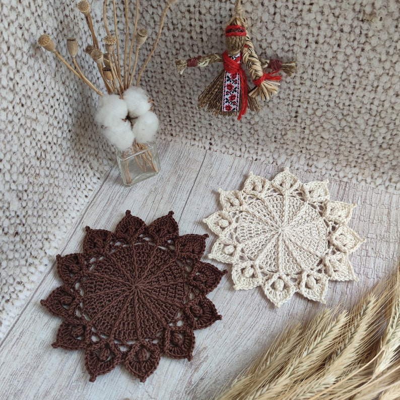 May include: Two crocheted doilies, one brown and one cream, with intricate patterns. The doilies are on a light wood surface.