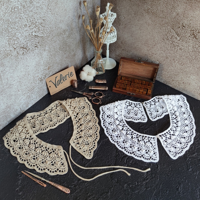 May include: Two crocheted collars, one beige and one white, with delicate lace patterns. The collars are laid out on a dark surface with vintage sewing supplies and a wooden box of letter stamps.