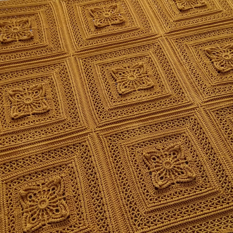 May include: Mustard yellow crocheted blanket with a repeating square pattern. Each square features a raised floral design in the center, surrounded by intricate openwork. The blanket is made of a soft, textured yarn.
