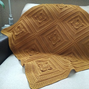 May include: Mustard-colored crocheted blanket with a repeating square pattern. Each square features a raised floral design. The blanket is draped over a light-colored sofa, showcasing its intricate texture and warm hue.