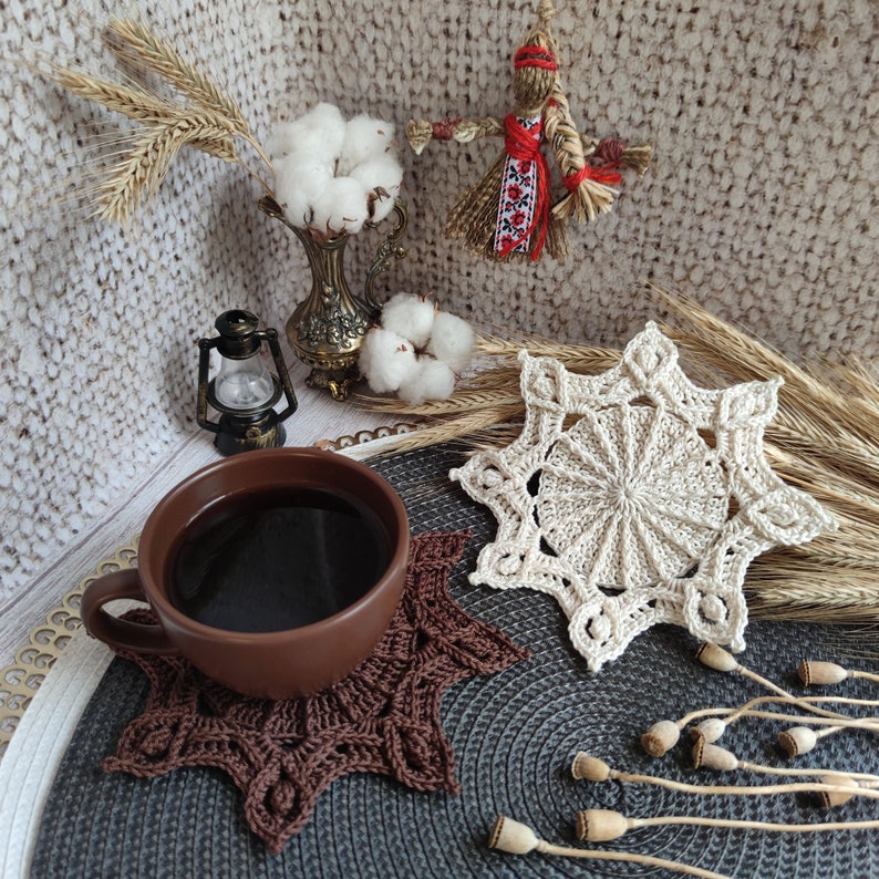 Puede incluir: Un posavasos de ganchillo marr&oacute;n con un posavasos de ganchillo beige encima. Los posavasos est&aacute;n sobre una superficie gris con una taza de caf&eacute; y una peque&ntilde;a linterna.