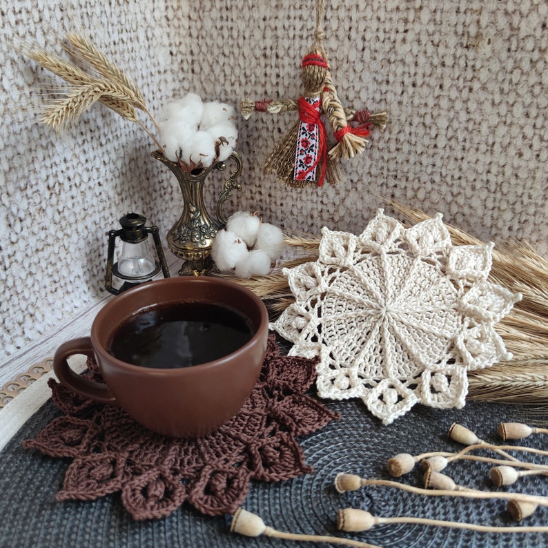 May include: A brown ceramic mug filled with dark coffee sits on a brown crocheted coaster. A white crocheted doily with a starburst design is in the background.