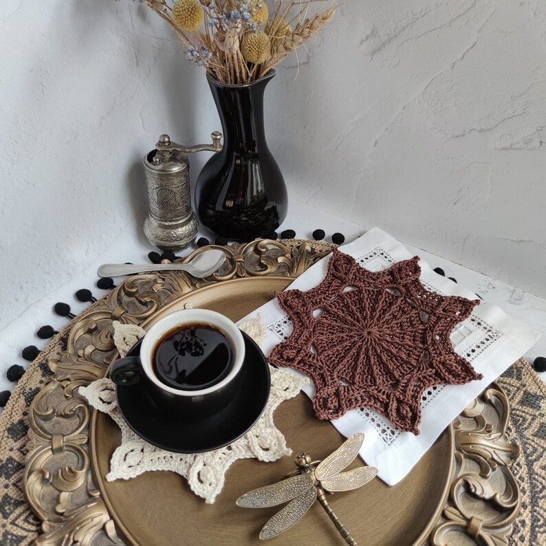 Puede incluir: Un posavasos de ganchillo marr&oacute;n con un dise&ntilde;o de estrella se encuentra sobre una servilleta blanca en una bandeja dorada y negra. Una taza de caf&eacute; se encuentra sobre un posavasos de ganchillo blanco junto al posavasos marr&oacute;n. Una figura de lib&eacute;lula est&aacute; en la bandeja.