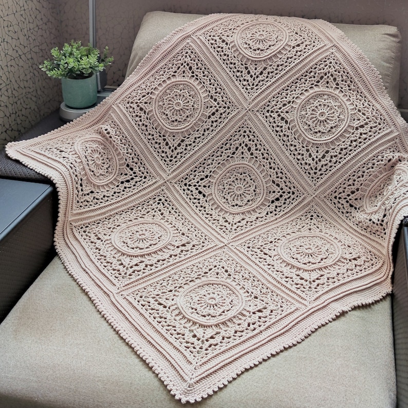 Pu&ograve; includere: Una coperta all'uncinetto di colore beige chiaro con un delicato e intricato motivo di cerchi e quadrati. La coperta &egrave; drappeggiata su un divano.