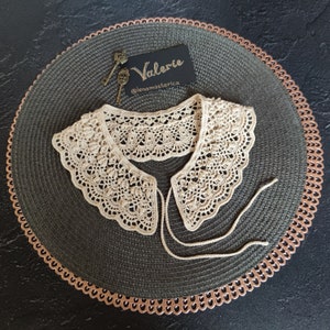 May include: A delicate crocheted collar in a cream color with a scalloped edge. The collar is laid flat on a dark green surface with a copper-colored border. A small card with the text "Valerie @lenamasterica" is visible in the background.