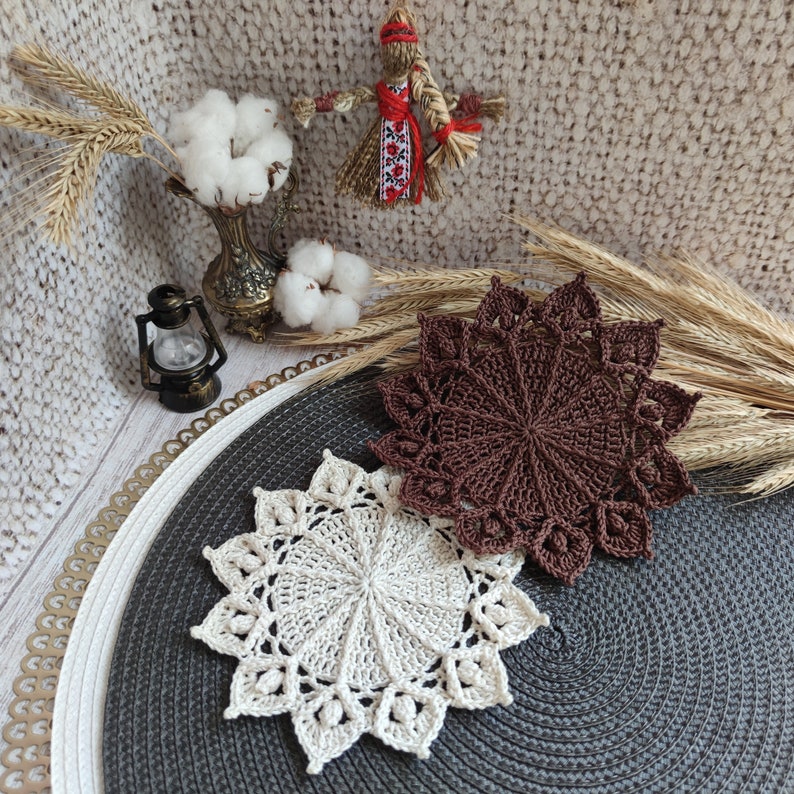 May include: Two crocheted doilies, one brown and one white, are placed on a dark grey round placemat. The doilies are both circular and have a delicate, intricate design.