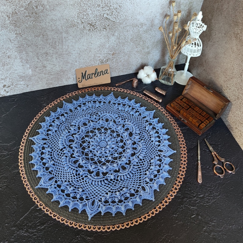 May include: A blue crocheted doily with a delicate floral pattern, displayed on a copper-colored round tray. The doily is a circular design with a central motif and intricate lacework.