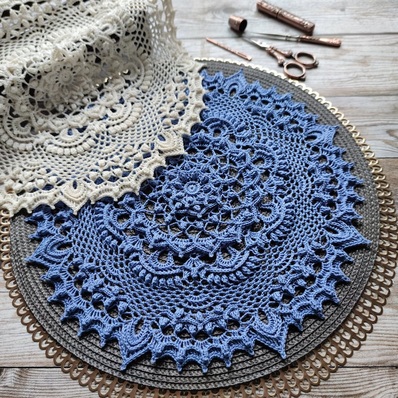 May include: Two crocheted doilies, one white and one blue, are displayed on a round, gold-trimmed, woven placemat. The blue doily is larger and has a more intricate design.