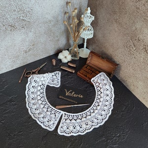 May include: White crocheted collar with a scalloped edge. The collar is laid flat on a black surface. The text "Valerie @lenamasterica" is on a white card.
