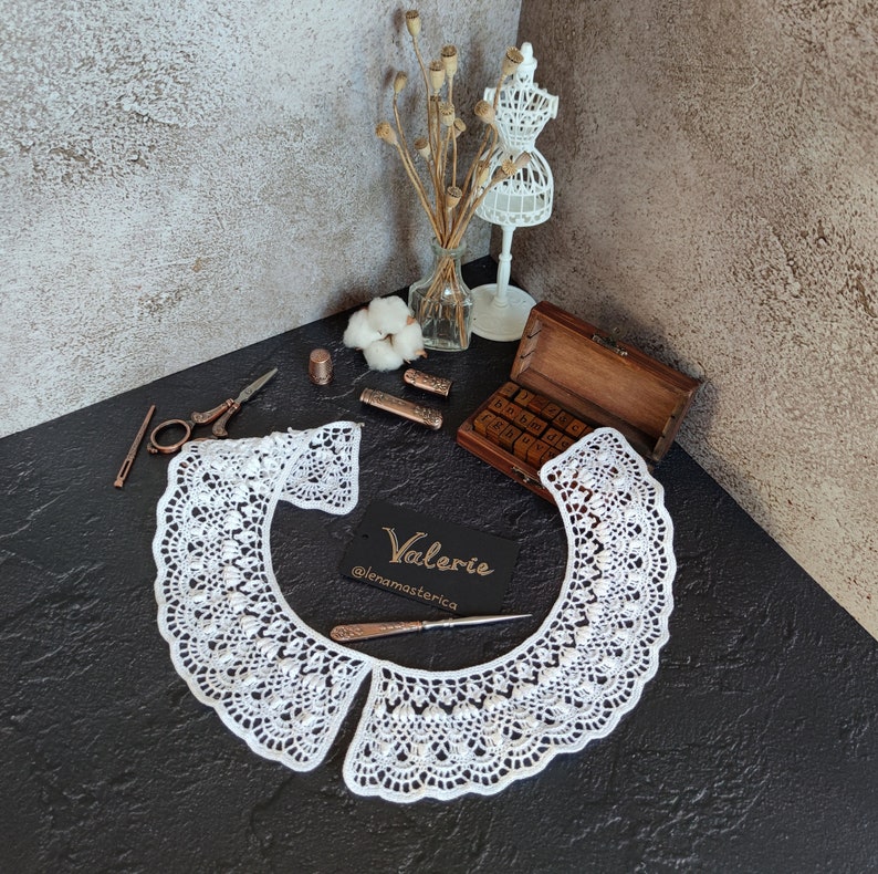 May include: White crocheted collar with a scalloped edge. The collar is laid flat on a black surface. The text "Valerie @lenamasterica" is on a white card.