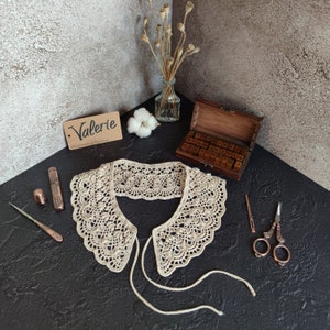May include: A crocheted lace collar with a delicate pattern. The collar is beige and has a tie closure. The collar is displayed on a dark surface with a pair of scissors, a needle, and a small wooden box with letters.