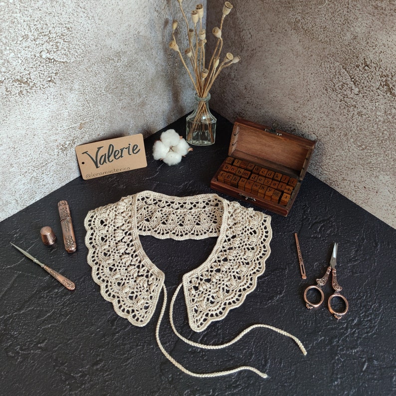 May include: A crocheted lace collar with a delicate pattern. The collar is beige and has a tie closure. The collar is displayed on a dark surface with a pair of scissors, a needle, and a small wooden box with letters.