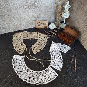 May include: Two crocheted collars, one beige and one white, with a scalloped edge. The collars are laid out on a black surface with a wooden box of letter stamps and other craft supplies.