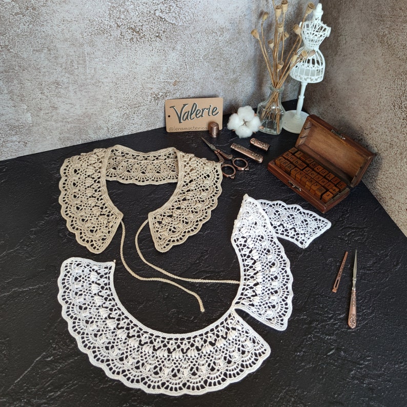 May include: Two crocheted collars, one beige and one white, with a scalloped edge. The collars are laid out on a black surface with a wooden box of letter stamps and other craft supplies.