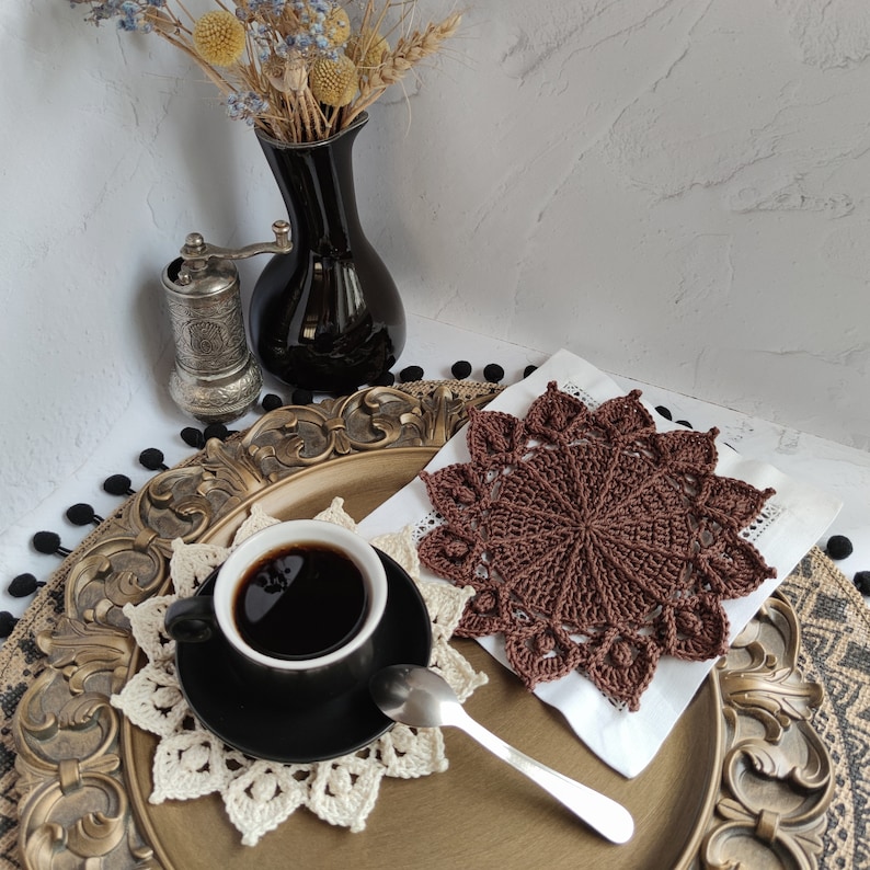 May include: A brown crocheted coaster with a decorative design sits on a white doily. A cup of coffee sits on the coaster. The coaster is on a gold-coloured tray with a black vase and dried flowers in the background.